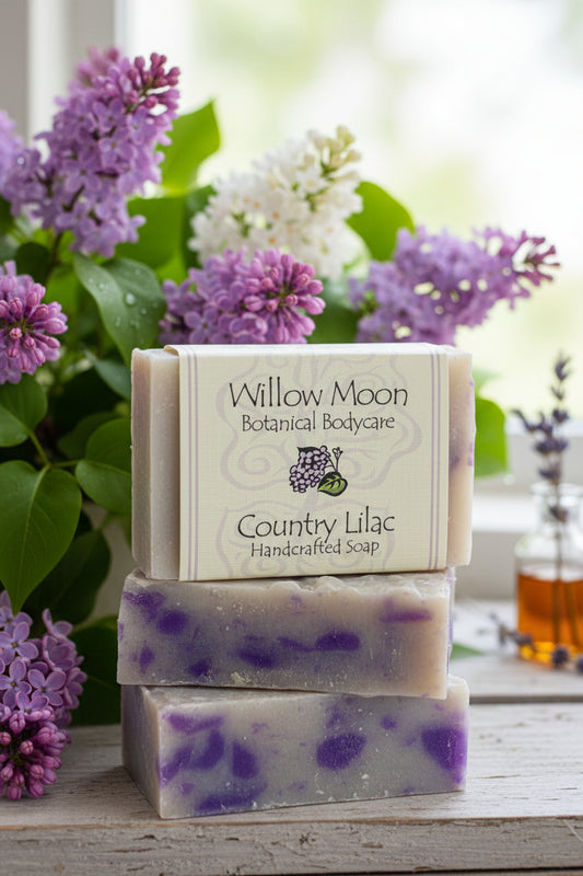 Three stacked bars of Willow Moon Botanical Bodycare 'Country Lilac' handmade soap, featuring a cream-colored label and purple marbled soap bars, styled with fresh lilac flowers on a rustic white wooden surface.
